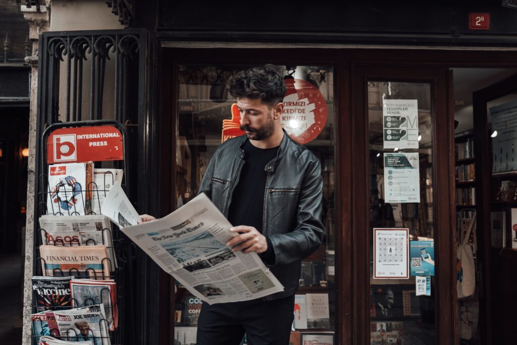 Serious bearded male with trendy haircut in casual clothes reading newspaper while standing near entrance of shop